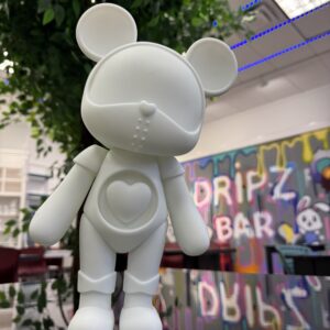 White toy figure with a heart emblem at Dripz Bar, promoting summer workshops.